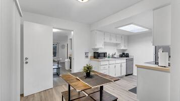 A modern kitchen with white cabinets, a microwave, and a coffee maker. There is a wooden table with a plant on it, and a bench.