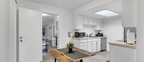 A modern kitchen with white cabinets, a microwave, and a coffee maker. There is a wooden table with a plant on it, and a bench.