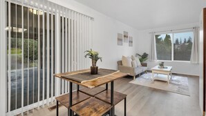 Una sala de estar moderna con una mesa de centro de madera, un sofá y una mesa de comedor. Hay plantas en macetas sobre la mesa de centro y vistas al exterior a través de amplios ventanales.