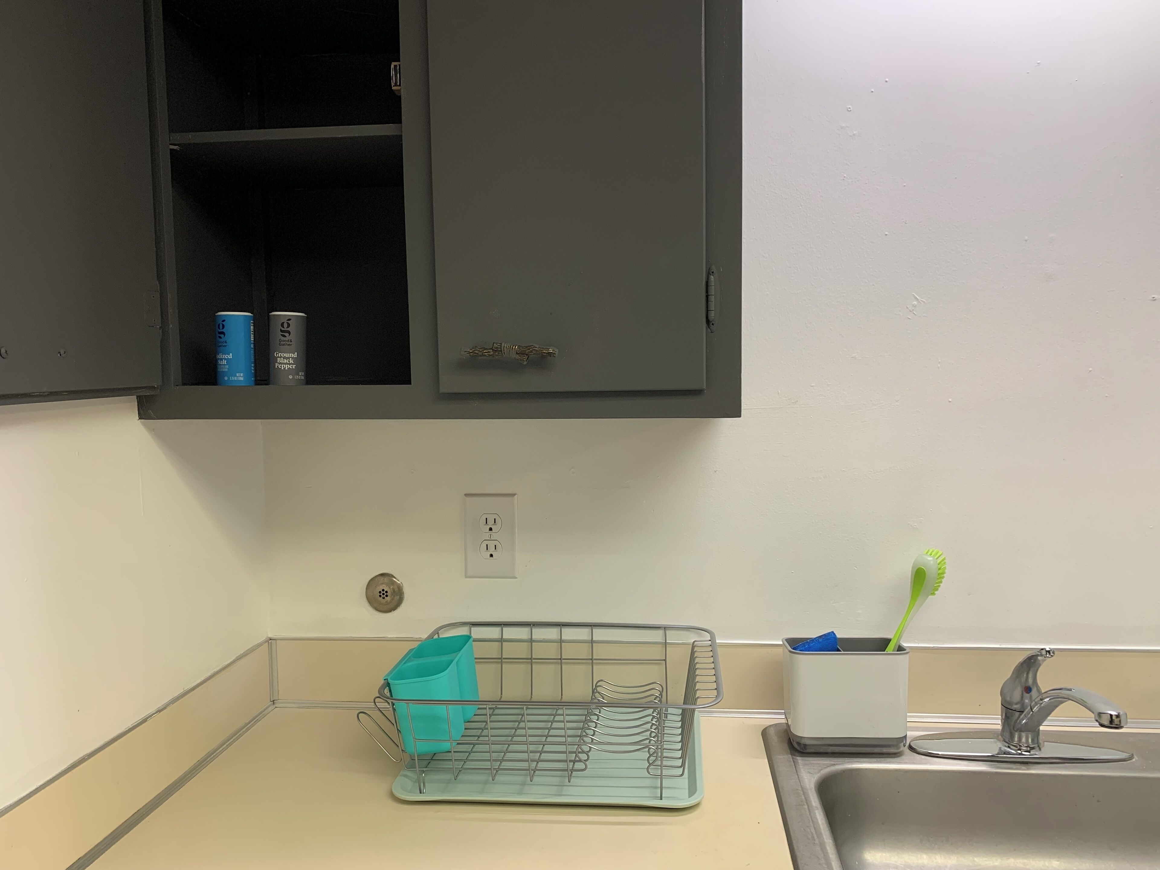 A kitchen with a sink, a countertop, a cabinet with a handle, and a metal rack with a blue container.