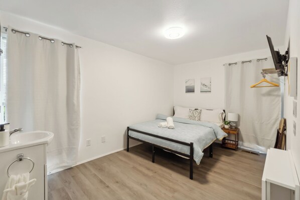 Une chambre à coucher comprenant un lit, une table de chevet, une lampe suspendue, une fenêtre avec des rideaux, et une salle de bain avec un lavabo et un miroir.
