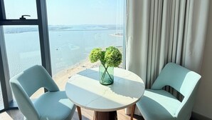 Una habitación moderna con una mesa redonda, dos sillas, un florero con flores verdes y vista al mar a través de amplios ventanales.