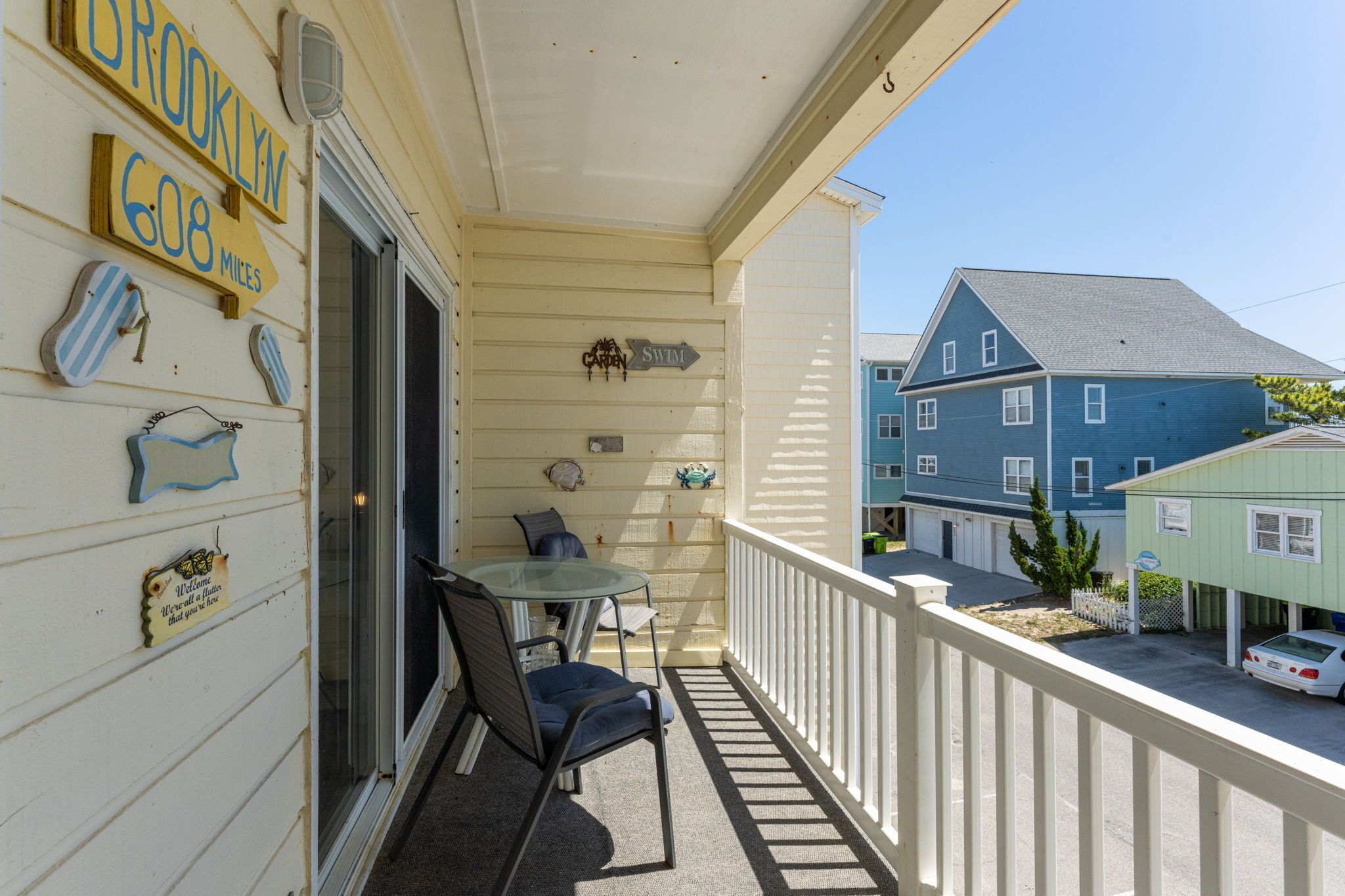 Ein Balkon mit Tisch und Stühlen, ein Schild mit der Aufschrift 'Brooklyn 608 Miles' und ein Blick auf ein blaues Haus im Hintergrund.