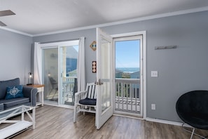 Ein Zimmer mit grauer Wand, einer weiĂen TĂŒr, die auf einen Balkon fĂŒhrt, einem grauen Sofa mit Kissen in Kompassoptik, einem weiĂen Stuhl und einem schwarzen Stuhl.