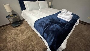 A neatly made bed with white linens and a dark blue throw, a bedside lamp, and a folded towel on the bed. - Convenient, Clean, Cozy, Cheap 2 (Tallahassee)