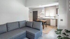 A modern living room with a grey sectional sofa, a dining area with a wooden table and chairs, and a kitchen with white appliances and a window with blinds. - NewOld Flat Bodrum Marina 2+1 (Bodrum)