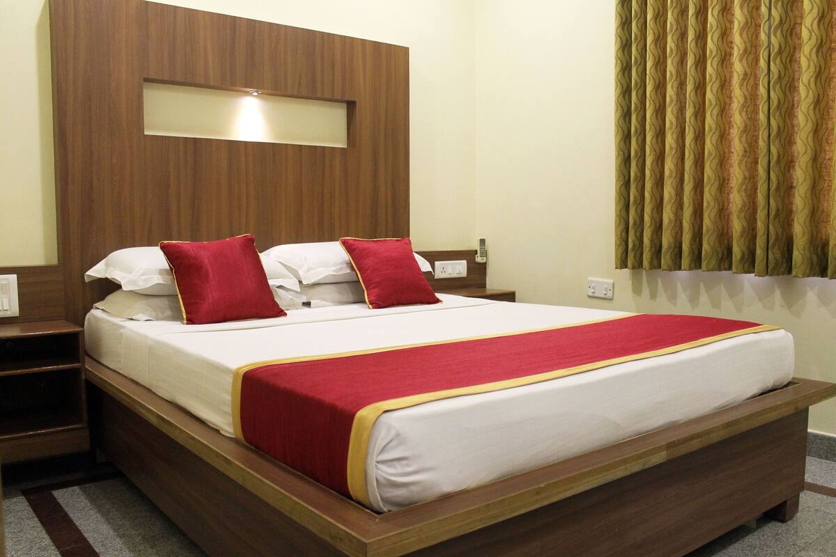 Une chambre d’hôtel avec une tête de lit en bois, un grand lit avec une literie blanche et des touches de rouge, une table de chevet et une fenêtre avec des rideaux.