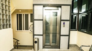 Um elevador em um corredor, com uma lixeira ao lado, um banco à direita e uma janela com persianas à esquerda.