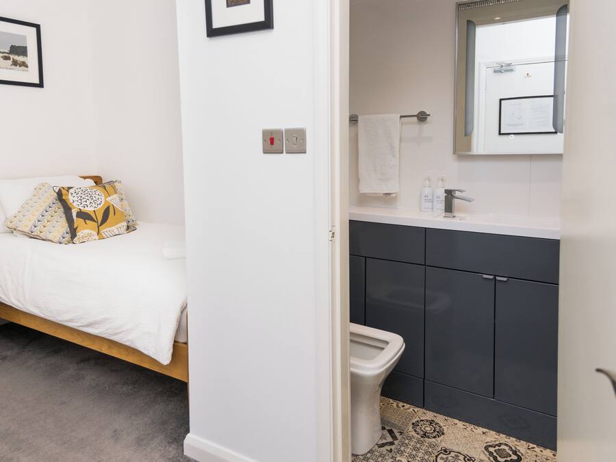 Un dormitorio con una cama, una mesita de noche y otra mesita auxiliar. Hay un baño con inodoro, lavabo y espejo.