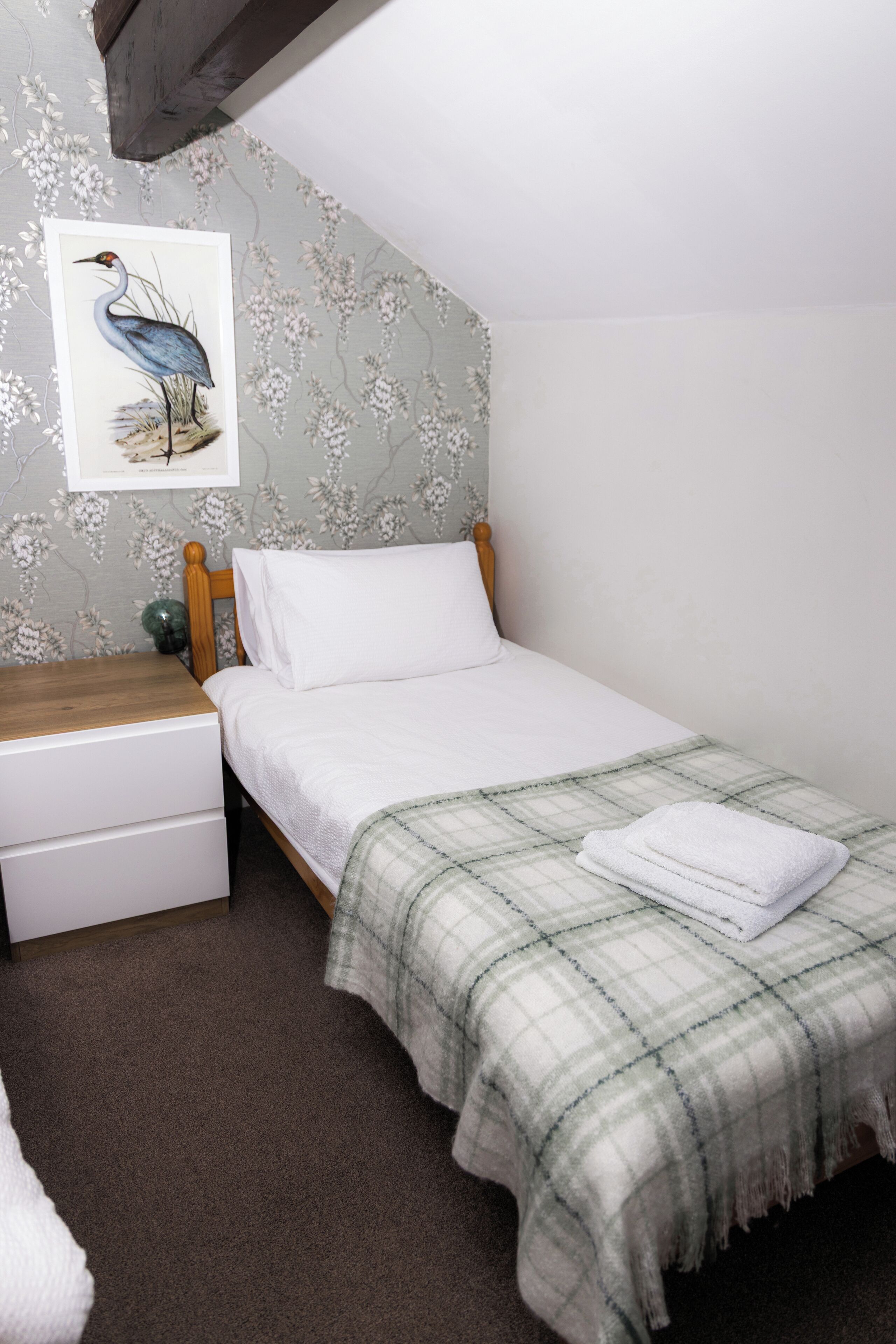 A single bed with a plaid blanket, a wooden headboard, a nightstand with a lamp, and a framed picture of a bird on the wall.