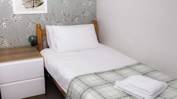 A single bed with a plaid blanket, a wooden headboard, a nightstand with a lamp, and a framed picture of a bird on the wall.