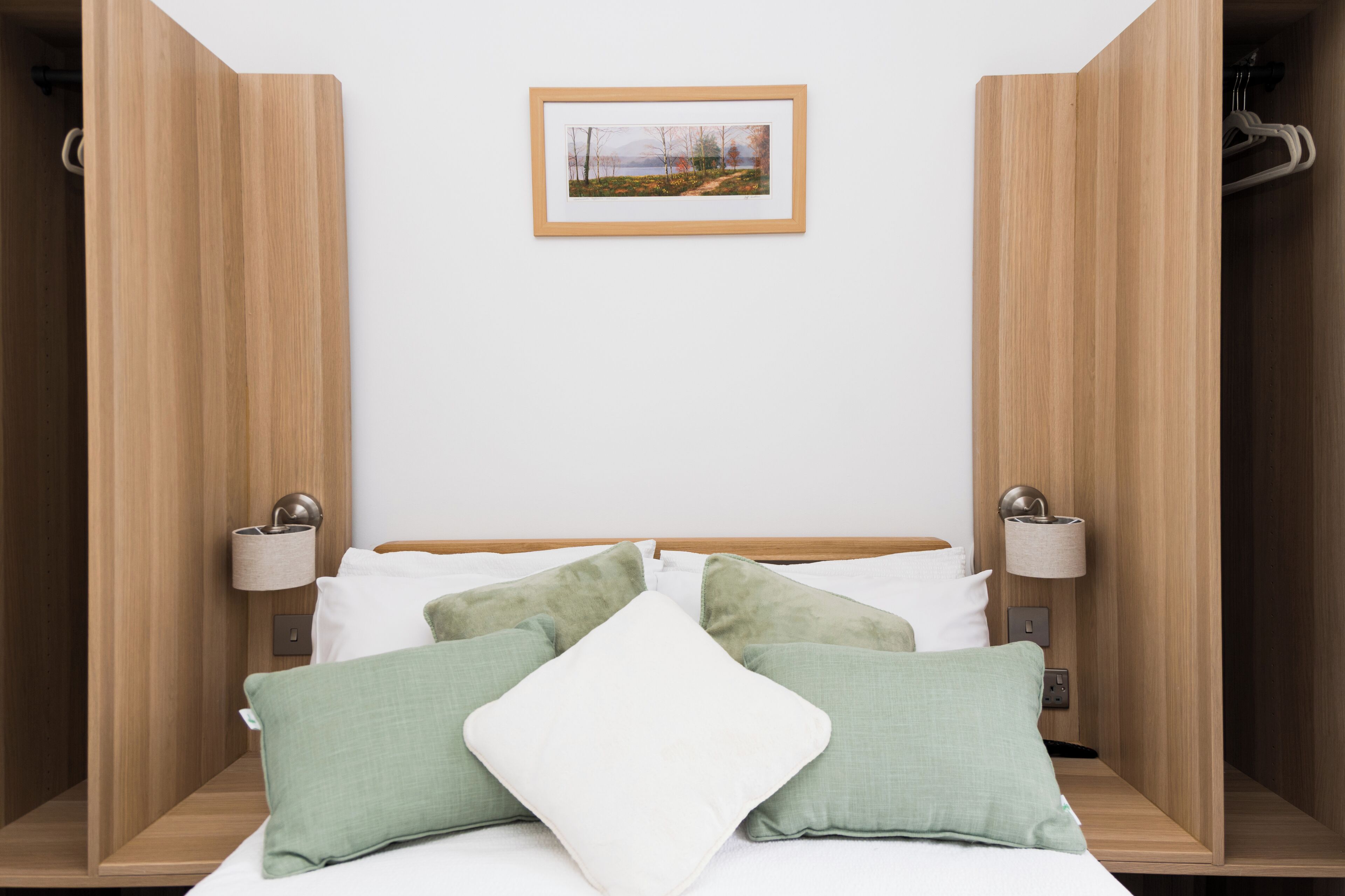 A neatly arranged bedroom with a bed, pillows, bedside lamps, and a framed picture on the wall.