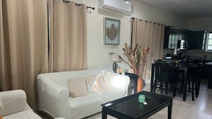 Ein Wohnzimmer mit einem weißen Sofa, einem schwarzen Couchtisch, beigen Vorhängen, einer Vase mit Trockenpflanzen und einer Küche im Hintergrund.