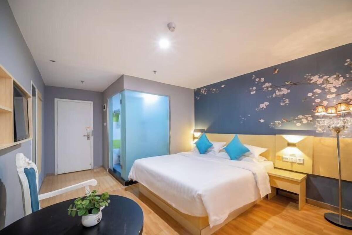A hotel room with a bed, bedside table, lamp, and a view of the outdoors.