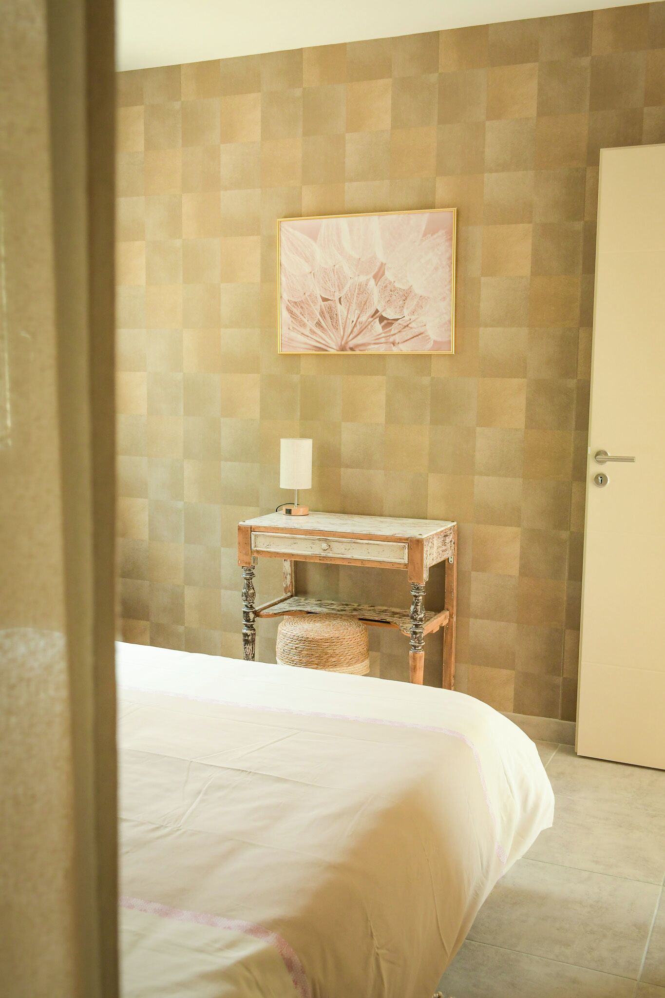 A hotel room with a bed, a small wooden nightstand with a lamp, a woven basket, and a framed picture on the wall.
