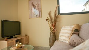 Una sala de estar con un televisor sobre un mueble de madera, una mesa de centro de vidrio con un cuenco decorativo, un sofá color beige con un cojín a rayas y un jarrón con plantas secas en la pared.