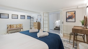 Ein Hotelzimmer mit einem großen Bett, einem Schreibtisch und einem Sessel. Es gibt eine Bank mit Kissen, eine Kommode und eine Tür, die zu einer Treppe führt.