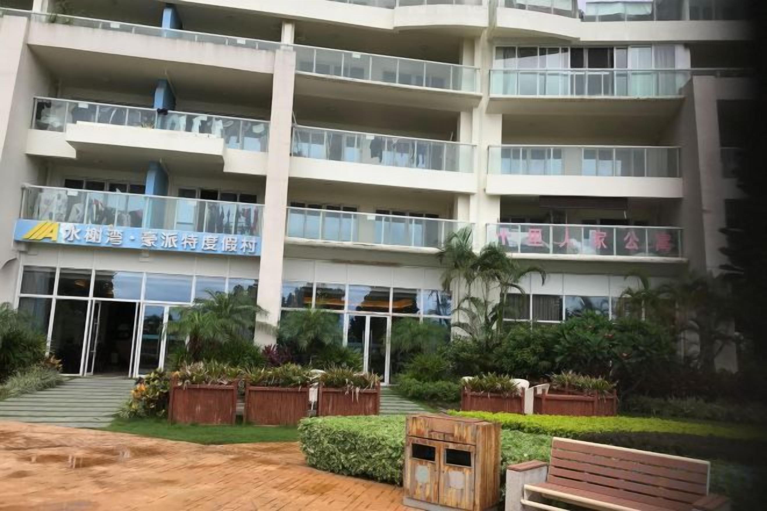 A multi-story building with balconies, a gated entrance, and a landscaped area with benches and plants.