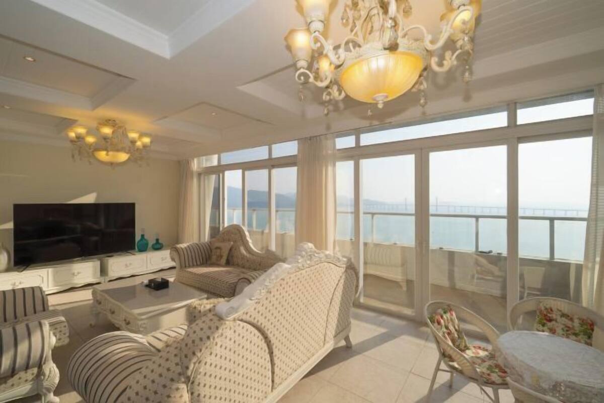 Amplia sala de estar con televisor grande, asientos cómodos, un balcón con vistas y un candelabro.
