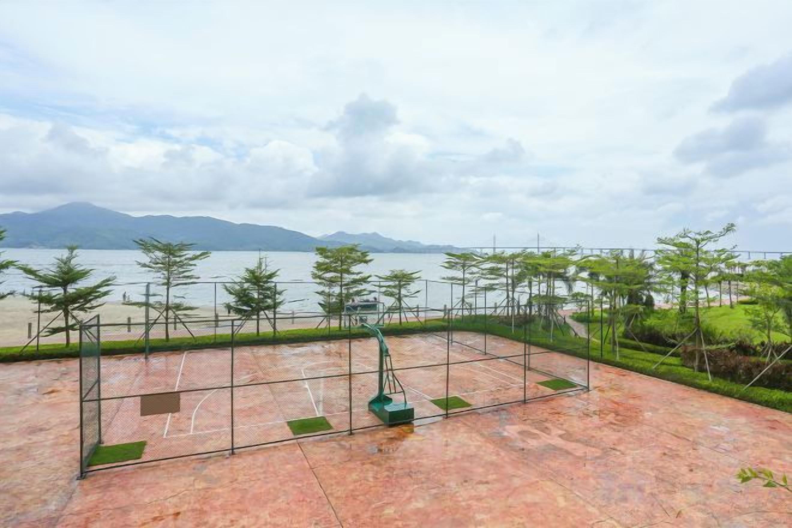 A tennis court with a net, surrounded by greenery and a view of a body of water.