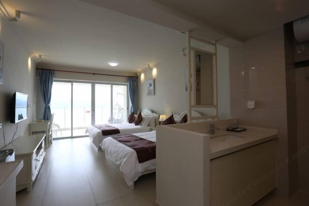 Habitación de hotel con dos camas, un ventanal grande con cortinas, un baño con espejo y un escritorio con silla.