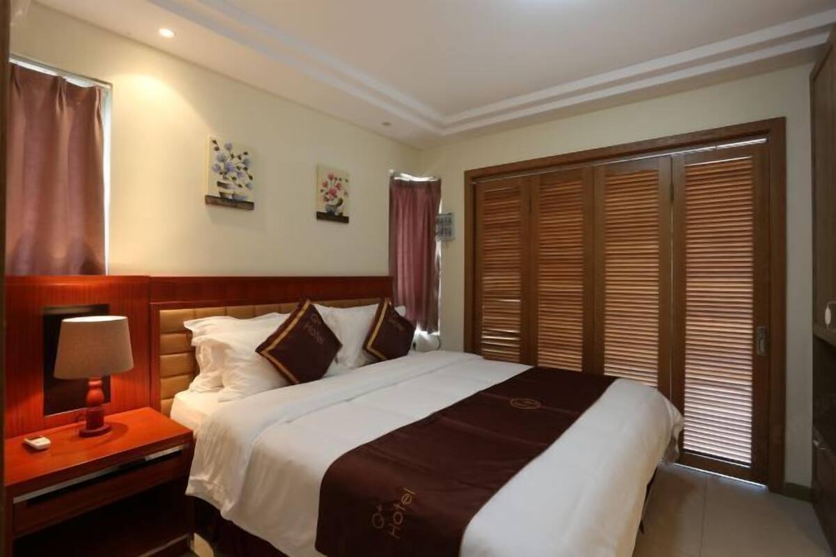 Una habitación de hotel con cama, mesita de noche, lámpara y un juego de contraventanas de madera.