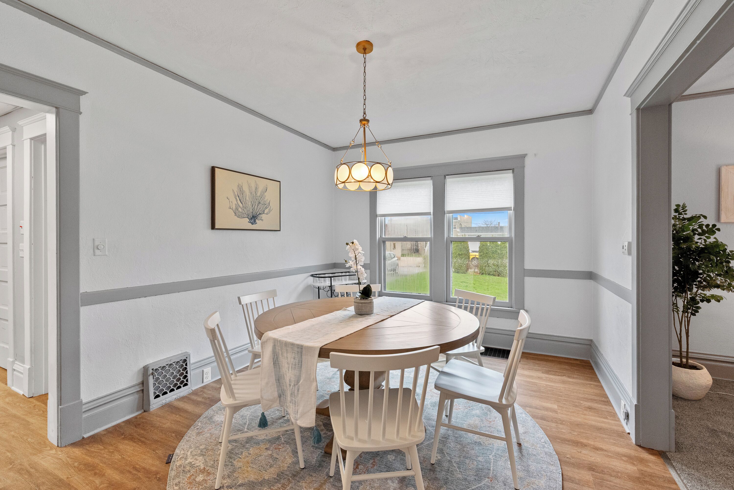 Een eetgedeelte met een ronde houten tafel, witte stoelen, een hanglamp en uitzicht door grote ramen.