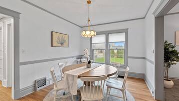 Een eetgedeelte met een ronde houten tafel, witte stoelen, een hanglamp en uitzicht door grote ramen.
