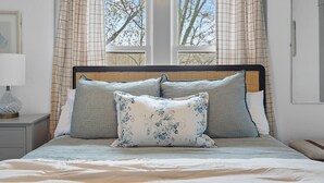 Ein ordentlich bezogenes Bett mit Kopfteil, ein Nachttisch mit Lampe und ein Fenster mit Vorhängen.