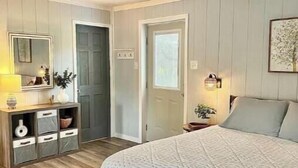 A bedroom with a large bed, a dresser with drawers, a mirror, a door, and a wall-mounted light fixture. - The Wren’s Nest (LaFollette)