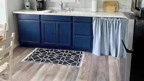A kitchen with white upper cabinets, a blue lower cabinet, a window, a sink, a cutting board, and a microwave. - The Wren’s Nest (LaFollette)