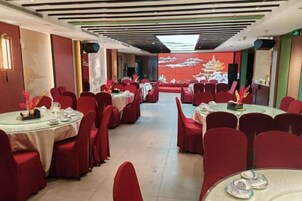 Un salón de banquetes con mesas redondas y sillas rojas, un escenario con un telón de fondo tradicional y una pantalla grande mostrando una escena festiva.