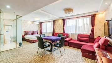 A hotel room with a bed, a dining table with chairs, a red sofa, a TV, and a bathroom with a glass partition.