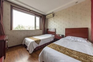 Habitación de hotel con dos camas individuales, suelo de madera, ventana con cortinas, unidad de aire acondicionado y un pequeño escritorio con teléfono.