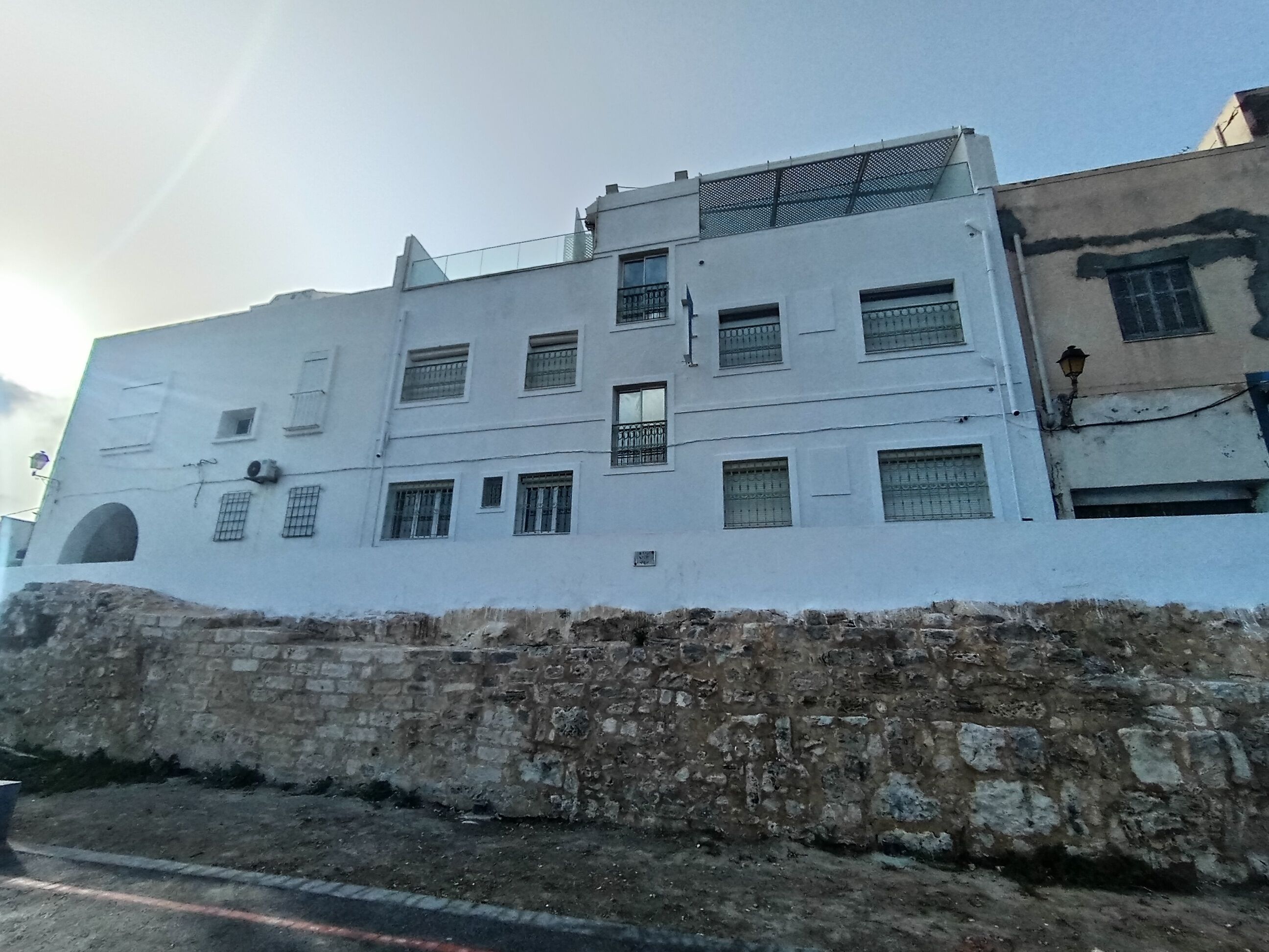 Un edificio de varios pisos con fachada blanca, múltiples ventanas con rejas de metal y una base de piedra.
