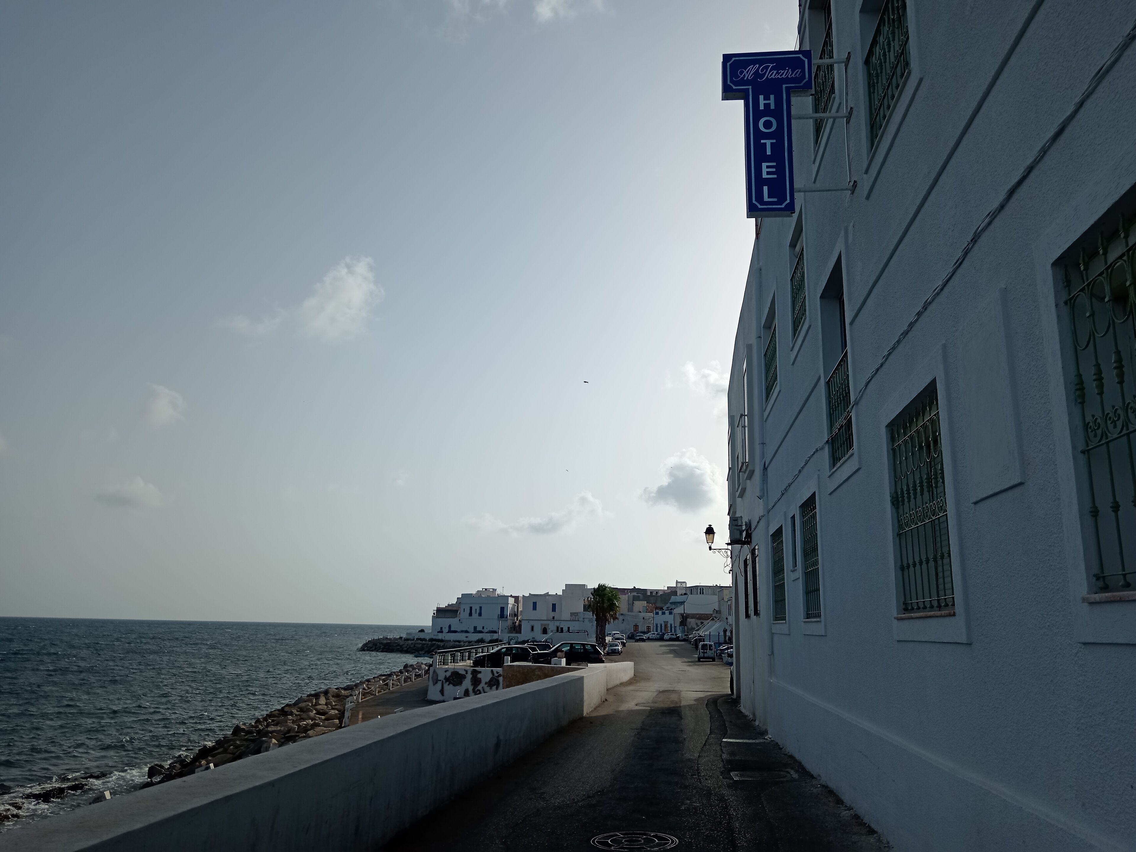 A coastal hotel with a sign indicating its name and type. The building has a white facade with multiple windows and a balcony. The view includes the sea and a rocky shoreline.