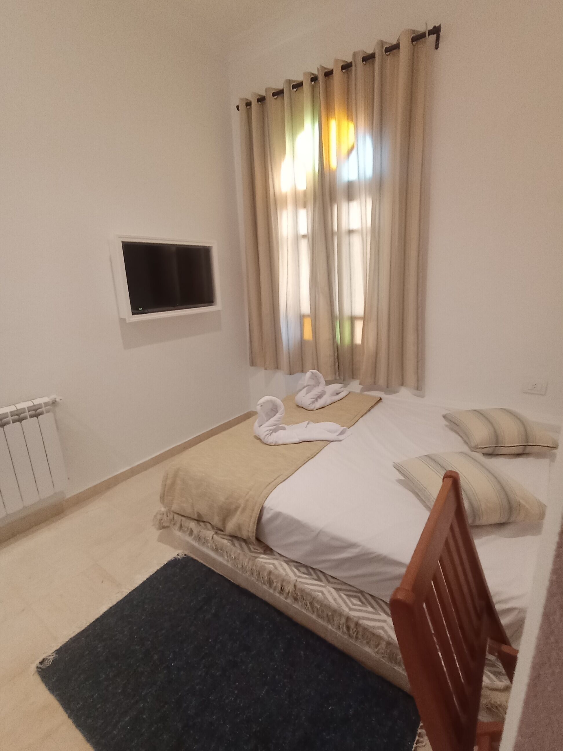 Ein ordentlich eingerichtetes Schlafzimmer mit einem Bett, einem an der Wand befestigten kleinen Fernseher und einem Fenster mit Vorhängen.