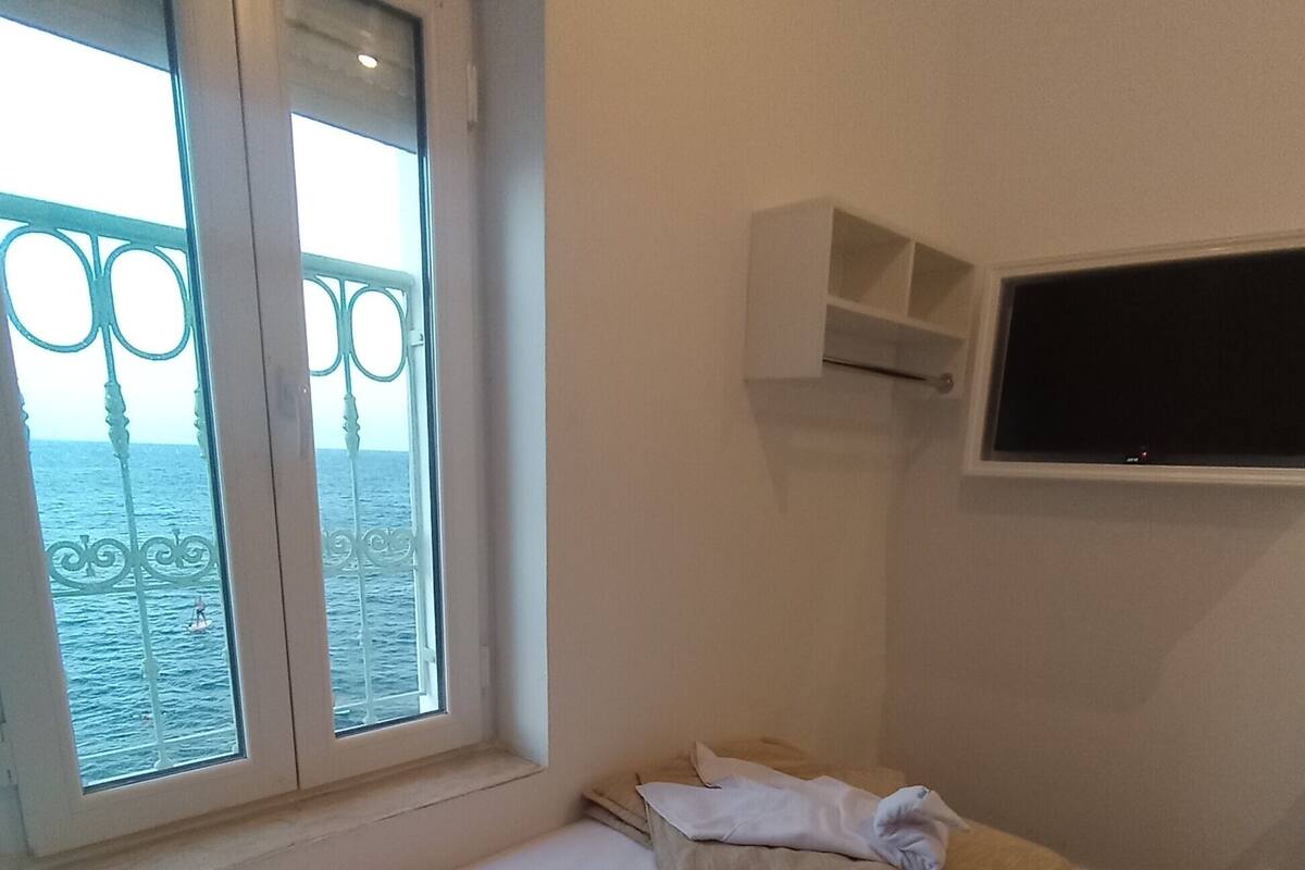 A small, simple room with a bed, a window with a view of the sea, and a mounted TV.