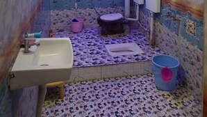 Ein Badezimmer mit Waschbecken, Toilette und Dusche. Die Wände haben ein strukturiertes Muster, und auf dem Boden stehen zwei Eimer.