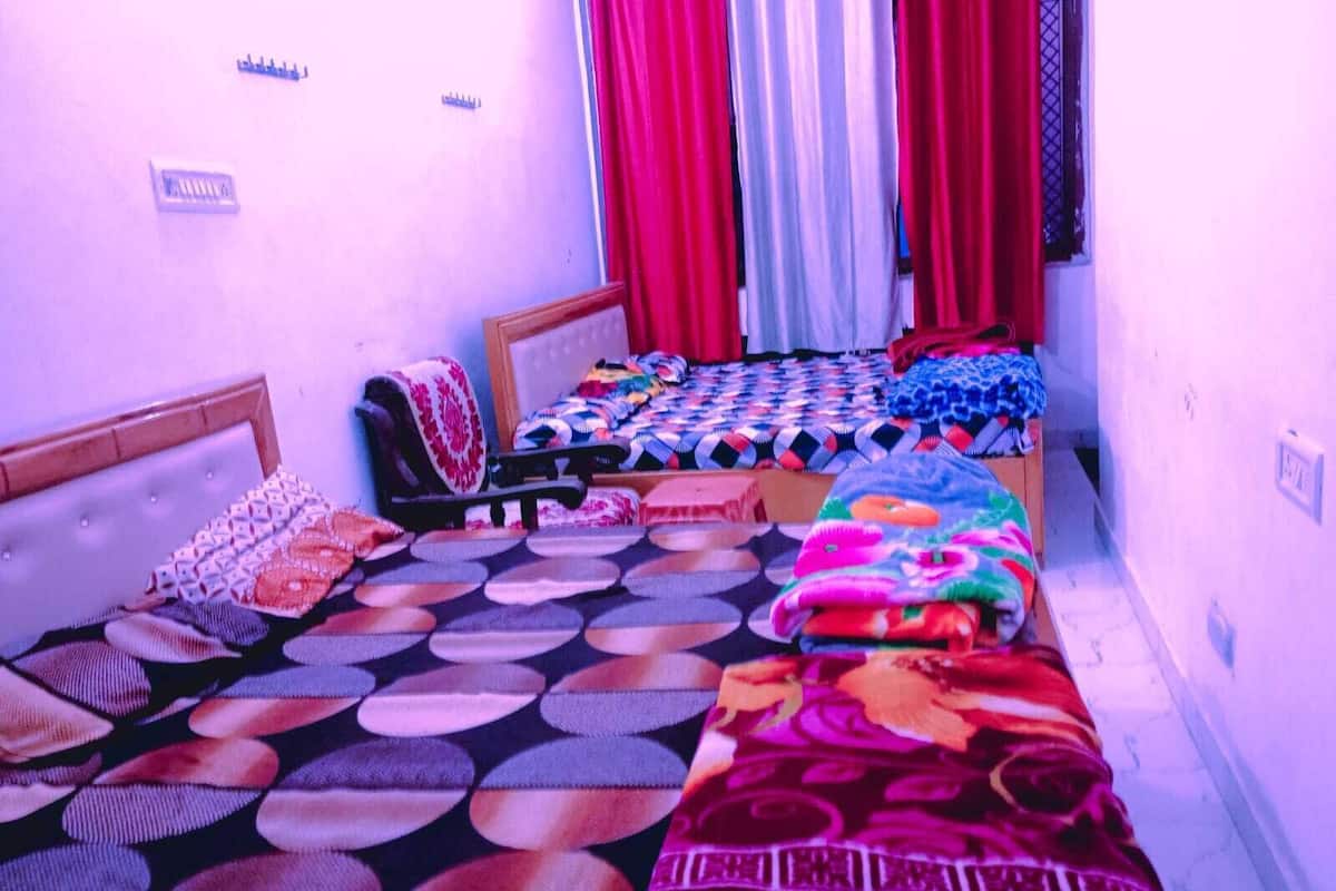 Una habitación pequeña y estrecha con dos camas, cada una con una colcha de diseño diferente. Hay cortinas en las ventanas y un pequeño estante con objetos en la pared.