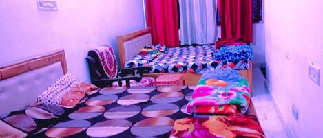 A small, narrow room with two beds, each with a distinct patterned bedspread. There are curtains on the windows, and a small shelf with items on the wall.