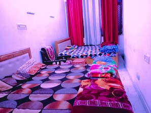 A small, narrow room with two beds, each with a distinct patterned bedspread. There are curtains on the windows, and a small shelf with items on the wall. - Shree mate guest house  (Faizabad)