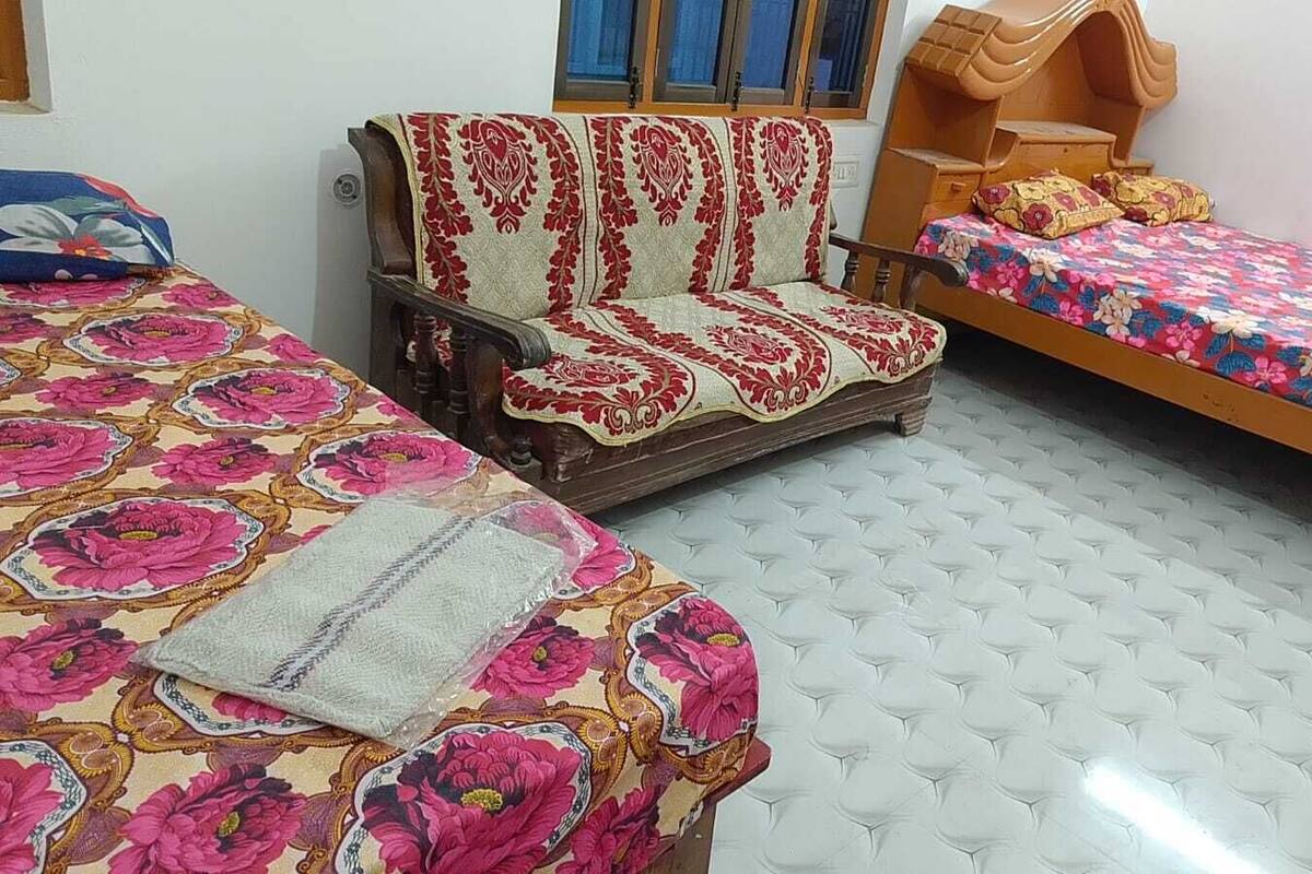 Una habitación con dos camas, una con una colcha estampada de flores y otra con un edredón estampado. Hay una silla de madera con funda estampada en rojo y blanco, y una cama con cabecera de madera.