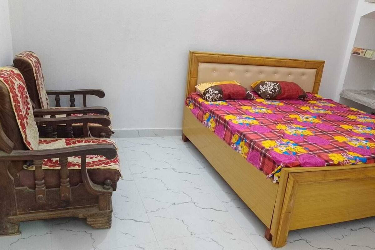 Una habitación con una cama de madera, una silla de madera con funda roja y blanca estampada, y un suelo de baldosas blancas.