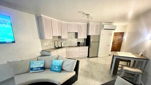 A modern kitchen with white cabinets, a built-in oven, and a refrigerator. There is a seating area with a sofa and cushions, and a small table with a chair.