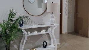 A white console table with a round mirror, a wall clock, a decorative lamp, and a dreamcatcher. There is a potted plant and a small turtle figurine on the floor.