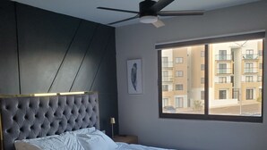 A bedroom with a tufted headboard, a large bed, a ceiling fan, a window with a view of buildings, and a framed picture on the wall.