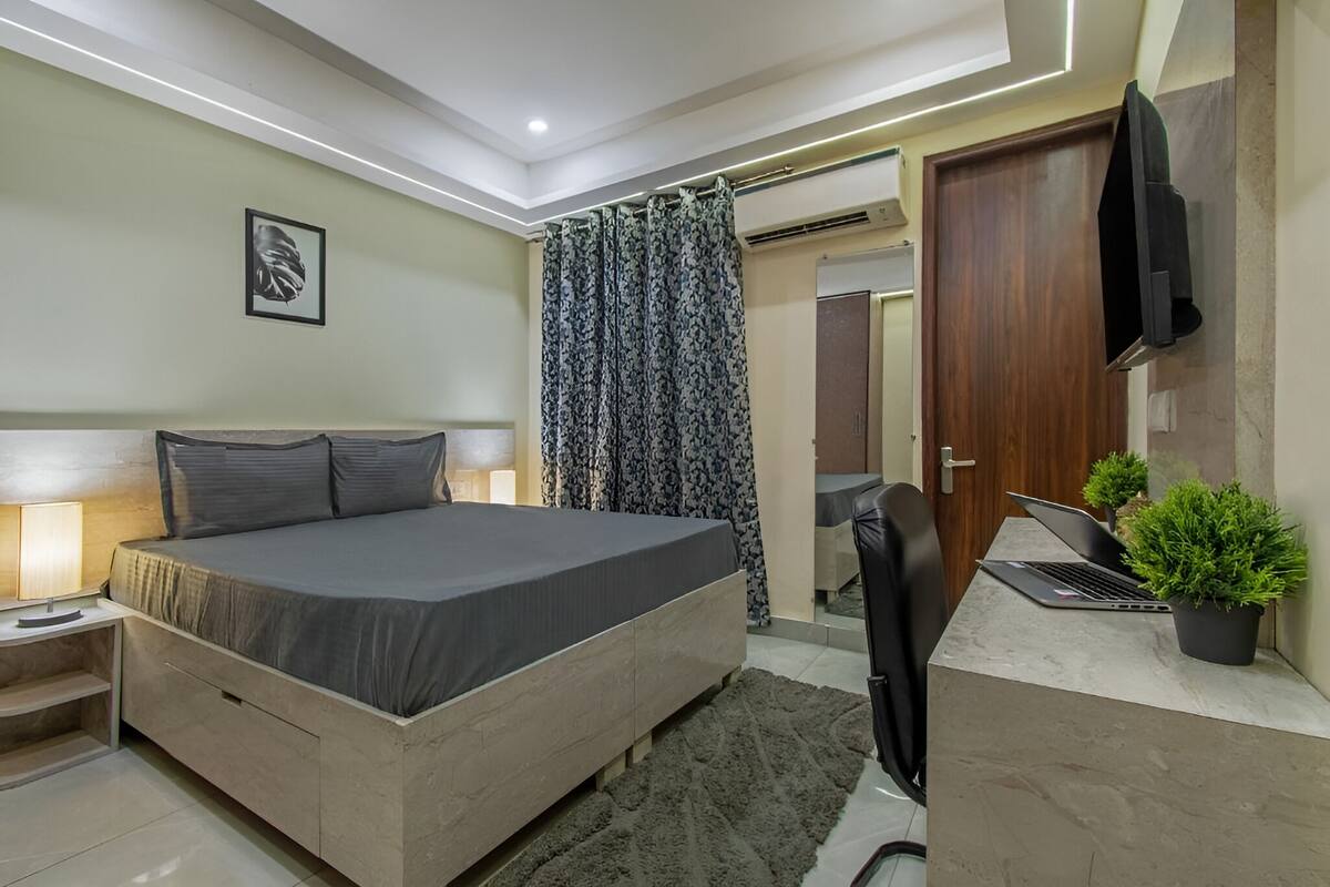 A modern bedroom with a bed, bedside table, lamp, and a laptop on a desk.