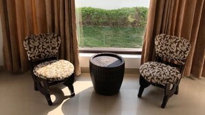 Une pièce comprenant deux fauteuils à motifs, un pouf rond en osier et une grande fenêtre avec des rideaux.