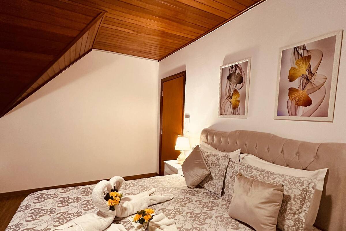Um quarto com teto inclinado, uma cama com roupa de cama estampada, duas mesas de cabeceira com abajures, e duas obras de arte florais emolduradas na parede.